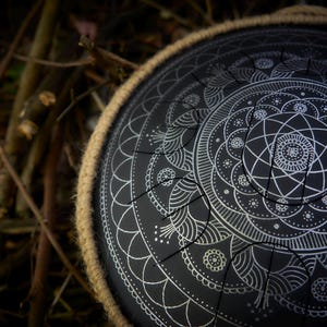 May include: A black steel handpan with a white mandala design. The handpan is surrounded by a brown leather strap.