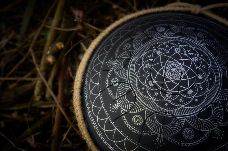 May include: A black steel handpan with a white mandala design. The handpan is surrounded by a brown leather strap.