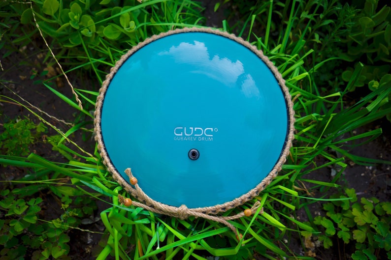 May include: A turquoise handpan with a rope handle and the text "GUD.C.O. GUAREV DRUM" on the surface.