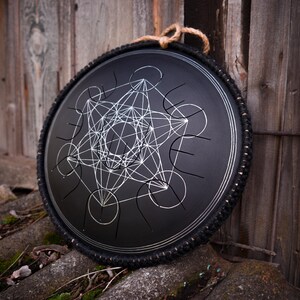 May include: A black handpan with a silver geometric design, surrounded by a black rubber band. The handpan is resting on a wooden surface.