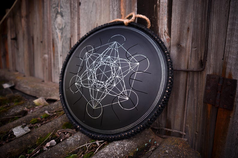 May include: A black handpan with a silver geometric design, surrounded by a black rubber band. The handpan is resting on a wooden surface.