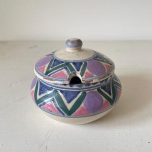 May include: A small, white ceramic sugar bowl with a lid. The bowl and lid are decorated with a geometric pattern in shades of pink, green, blue, and purple.
