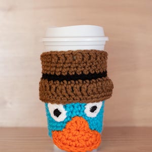 May include: A handmade crochet coffee cup cosy featuring a cartoon platypus design. The cosy is brown with a black stripe, blue face, orange beak, and white eyes. The cup has a white lid.