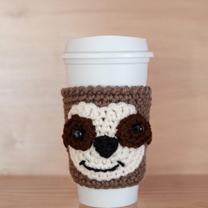 May include: A white coffee cup with a brown and white crocheted sloth face cosy. The sloth has brown eyes and a black nose.
