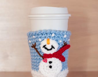 May include: A blue and white crocheted coffee cup cozy with a snowman design. The snowman is wearing a red scarf and has a small red heart on its chest.