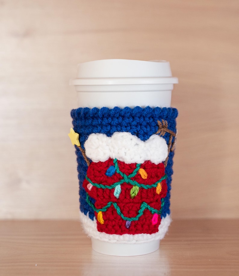 May include: A blue and red crocheted coffee cup cozy with a white snow-capped Christmas tree design and colorful lights.
