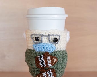 May include: A crocheted coffee cup cozy in the shape of a person wearing glasses and a face mask. The cozy is green, white, and brown with a pattern of footballs.