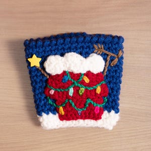 May include: A blue crocheted coffee cup cozy with a white crocheted snow cap and a red crocheted Christmas tree with green garland and colorful lights.