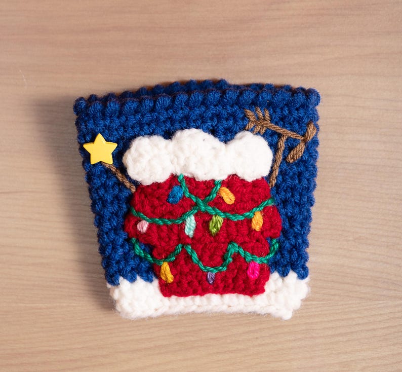 May include: A blue crocheted coffee cup cozy with a white crocheted snow cap and a red crocheted Christmas tree with green garland and colorful lights.