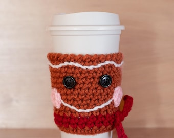 May include: A white to-go cup with a crocheted gingerbread man cozy. The cozy is brown with white icing details and black button eyes. It has a red crocheted scarf.