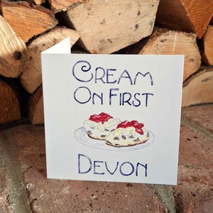 May include: A white card with blue text that reads "Cream on First Devon". The card features a hand-drawn illustration of two scones with jam and cream on a white plate.