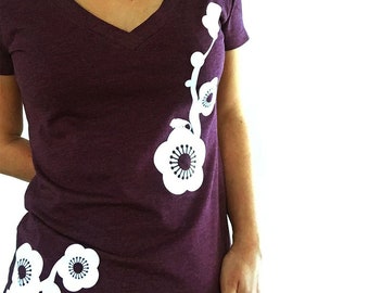 May include: A purple v-neck t-shirt with a white floral design. The design features a stylized branch with flowers and leaves.