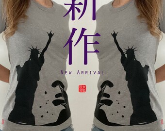 May include: A gray t-shirt with a black silhouette of the Statue of Liberty and a Japanese wave design. The shirt has the text "新作" in purple on the front. The text "NEW ARRIVAL" is below the Japanese characters. A red stamp with the text "百紋" is below the text.