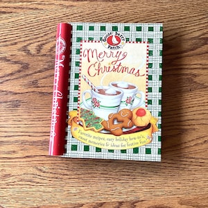 May include: A cookbook titled "Merry Christmas" with a red spine and a green and white checkered cover. The cover features illustrations of hot chocolate, cookies, and the text "Merry Christmas". The book is on a wooden surface.