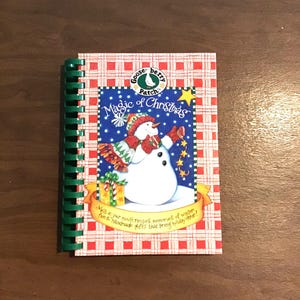 May include: A spiral-bound cookbook titled Magic of Christmas by Gooseberry Patch. The cover features a red and white checkered pattern, a central illustration of a snowman on a blue background with stars, and text reading Melt-in-your-mouth recipes, memories of winter, fun & handmade gifts that bring holiday cheer!