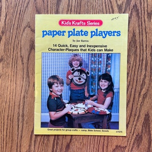 May include: A vintage book titled "Paper Plate Players" by Joe Barros, part of the Kid's Krafts Series. The cover features three children crafting paper plate characters. The book promises 14 quick and easy character-plaques for kids to make.