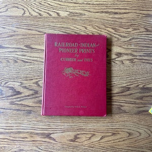 以下が含まれることがあります： 「Railroad-Indian and Pioneer Prints by Currier and Ives」というタイトルの赤いハードカバーの本。本は木製のテーブルの上に置かれています。