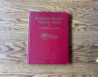 May include: A red hardcover book titled "Railroad-Indian and Pioneer Prints by Currier and Ives". The book is on a wooden table.