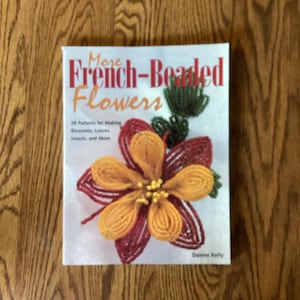 Puede incluir: Un libro titulado "More French-Beaded Flowers" con un diseño de flores con cuentas en naranja, rojo y verde. La portada incluye texto sobre patrones para flores, hojas, insectos y más. El libro está sobre una superficie de madera.