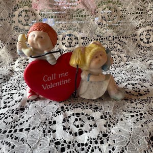 Puede incluir: Figura de cerámica de San Valentín con dos figuras de querubines. Uno sostiene un teléfono, el otro está sentado junto a un corazón rojo con la inscripción "Call me Valentine". Las figuras tienen pelo rubio y rojo, y visten colores pastel.