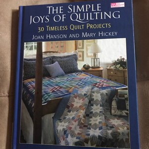 May include: The Simple Joys of Quilting book cover featuring a blue and white quilt on a bed with a wooden headboard. The book title is "The Simple Joys of Quilting" with the subtitle "30 Timeless Quilt Projects". The authors are Joan Hanson and Mary Hickey.