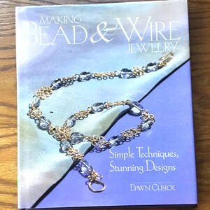 May include: A softcover book titled Making Bead & Wire Jewelry by Dawn Cusick. The cover features a blue and green watercolor background with a photograph of a gold wire and blue bead necklace. The text reads Simple Techniques, Stunning Designs.