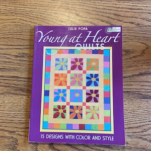 Puede incluir: Un libro titulado "Young at Heart Quilts" de Julie Popa, con un diseño de edredón colorido en la portada. El diseño del edredón incluye patrones florales en varios colores. La portada del libro es morada.