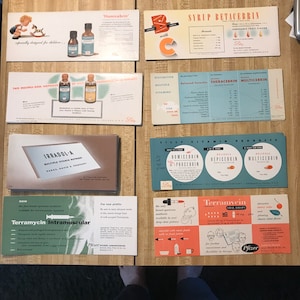May include: A collection of vintage pharmaceutical advertisements and product packaging. The items feature colorful graphics and text, including "Homicebrin," "Syrup Betacebrin," and "Terramycin." The ads showcase illustrations and product information.