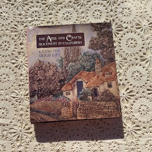 May include: A book titled "The Arts and Crafts Movement in California" with the subtitle "Living the Good Life." The cover shows a detailed illustration of a cottage with a thatched roof, surrounded by trees and a stone wall. The book is hardcover.