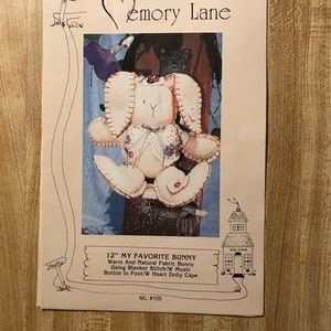 Può includere: Un modello di carta intitolato "Down Emory Lane" con un coniglietto di stoffa. Il coniglietto è color crema con dettagli cuciti, un colletto floreale e accenti a forma di cuore. Il testo recita "12" MY FAVORITE BUNNY".