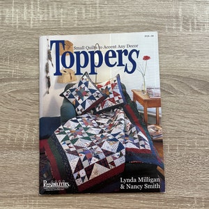Könnte beinhalten: Ein Buch mit dem Titel "Toppers" und dem Untertitel "Small Quilts to Accent Any Decor" wird auf einer Holzoberfläche angezeigt. Das Cover zeigt eine Steppdecke und Kissen mit Sternmustern. Die Autoren des Buches sind Lynda Milligan & Nancy Smith.