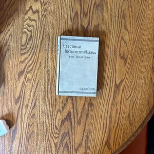 Puede incluir: Un pequeño libro antiguo titulado "Electrical Instrument-Making for Amateurs" de S.R. Bottone. El libro tiene una cubierta azul claro con texto negro y está sobre una mesa de madera con un acabado marrón cálido.