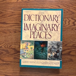 Puede incluir: Un libro de tapa dura titulado "The Dictionary of Imaginary Places" con una edición ampliada. La portada presenta ilustraciones y los nombres de los autores, Alberto Manguel & Gianni Guadalupi. El libro está sobre una superficie de madera.