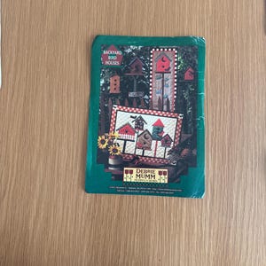 Puede incluir: Un libro con borde verde titulado "Backyard Bird Houses" con imágenes de casas para pájaros y un edredón con diseños de casas para pájaros. La portada también muestra el nombre "Debbie Mumm" y una dirección web.