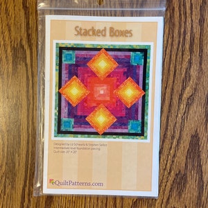 May include: A clear plastic bag containing a quilt pattern titled "Stacked Boxes." The pattern features a colorful geometric design with squares and rectangles in shades of orange, pink, blue, and green. The quilt size is 20" x 20".