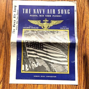 May include: A vintage sheet music cover titled The Navy Air Song, Pilots, Man Your Planes! The blue cover features a gold winged emblem, a black and white photograph of a large American flag on a ship deck, and text by Arthur Kurlan and Peter De Rose.