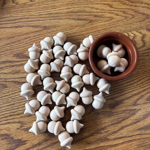May include: A collection of small, unfinished wooden acorn-shaped figurines. Some acorns are scattered on a wooden surface, while others are in a small, brown ceramic bowl. The acorns are light brown and have a natural wood grain.