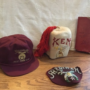 May include: A collection of vintage fraternal items, including a maroon baseball cap with gold embroidery reading "Medinah Juliet Shrine Club," a small red book, a fez, and a decorative block with "KEM" text.