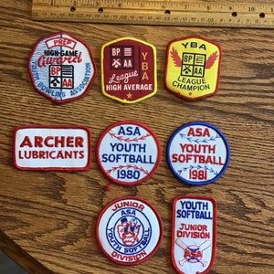 Puede incluir: Una colección de ocho parches bordados, la mayoría con diseños de béisbol. Los parches son rojos, blancos, azules y amarillos. Algunos de los parches tienen texto que dice "ASA Youth Softball", "YBA League Champion", "Prep High-Game Award" y "Archer Lubricants".