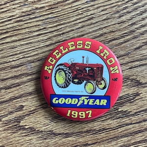 Puede incluir: Un botón redondo rojo con las palabras "AGELESS IRON" en amarillo. El botón presenta un tractor rojo con ruedas amarillas y el texto "GOOD YEAR 1997". El tractor está etiquetado como "1950 Massey-Harris 44".