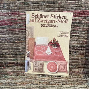 May include: A book titled "Schöner Sticken auf Zweigart-Stoff" with illustrations of cross-stitch designs, including a tablecloth, pillows, and a decorative dog statue. The book is on a multi-colored woven rug.