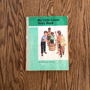 May include: A vintage children's book titled "My Little Green Story Book" with a green and white cover. The cover features illustrations of children and a basket of apples. The book is on a wooden surface.
