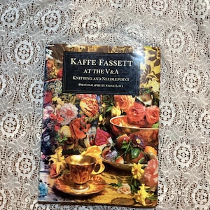 May include: A book titled "Kaffe Fassett at the V&A Knitting and Needlepoint" with a colorful floral cover. The cover features a gold-rimmed teacup and saucer, and a bowl of fruit. The book is on a white lace tablecloth.