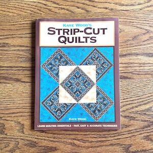 Puede incluir: Un libro de instrucciones de tapa blanda titulado Kaye Wood's Strip-Cut Quilts. La portada presenta un patrón de edredón geométrico en azul y blanco, bordes marrones y el texto: Learn Quilting Essentials - Fast, Easy & Accurate Techniques.
