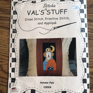 Könnte beinhalten: Ein Kreuzstichmuster von Val's Stuff mit dem Titel "Autumn Pals". Das Muster zeigt ein herbstliches Design mit einer Vogelscheuche, einer schwarzen Katze und einem Mond. Der Hintergrund ist cremefarben mit einem schwarz-weiß karierten Rand.