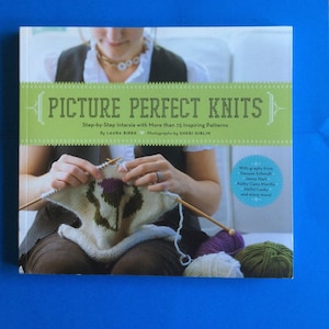 May include: A book titled "Picture Perfect Knits" with a green banner across the top. The book shows a person knitting a white and beige piece with a purple and green design. Knitting needles and yarn are visible.