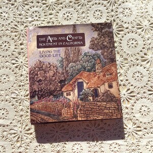 Może przedstawiać: Książka w twardej oprawie zatytułowana The Arts and Crafts Movement in California: Living the Good Life. Okładka przedstawia teksturowaną ilustrację kamiennego domku z dachem krytym strzechą, otoczonego drzewami i kamiennym murem w stylu malarskim.