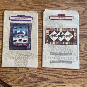 May include: Two brown paper bags with quilt patterns printed on them. The left bag has a moose, bears, and fish pattern with the text "Moose Junction 31" x 41". The right bag has a horse pattern with the text "Roundup 39" x 29". Both bags have the text "Quilt Designs by PINE NEEDLES McKenna Ryan".