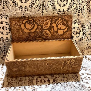 May include: Wooden box with a hinged lid, featuring carved rose designs. The box is rectangular with a light brown interior and a darker, textured exterior. The lid and front of the box are decorated with floral carvings.