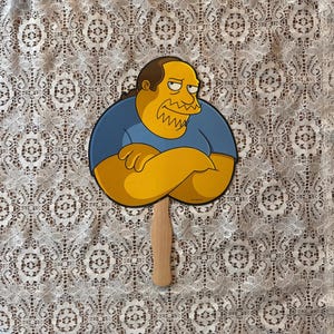 Op de afbeelding: Een handventilator met een stripfiguur met gele huid, een blauw shirt en gekruiste armen. De ventilator heeft een houten handvat en staat tegen een achtergrond met patronen.